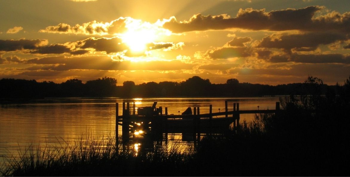 The sun rises over still water. There are long, scattered clouds across the sky with gold tones and a shining sun. The silhouette of a boat, dock, and marsh grass are in the foreground.
