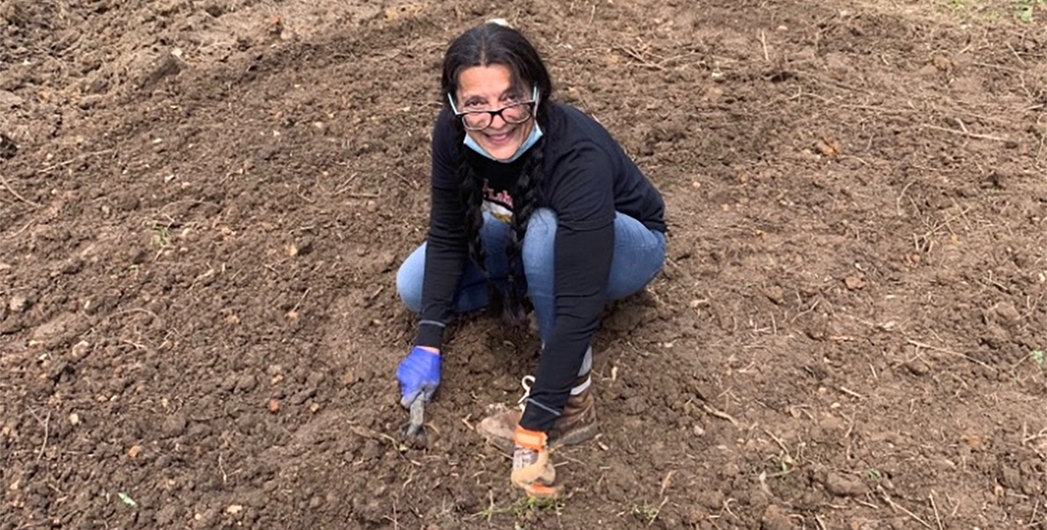A woman holding a spade crouches down in a dirt plot.