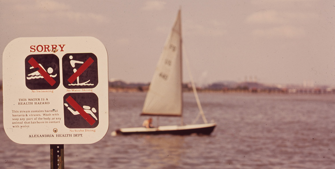In the background a main sails a small sailboat, while a sign from the Alexandria Health Department in the foreground bears the headline SORRY and images depicting No Swimming, No Water Skiing, and No Scuba Diving.