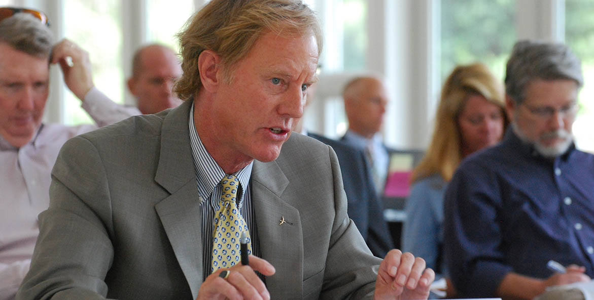 Man at a table speaking with audience and member of the press behind him.