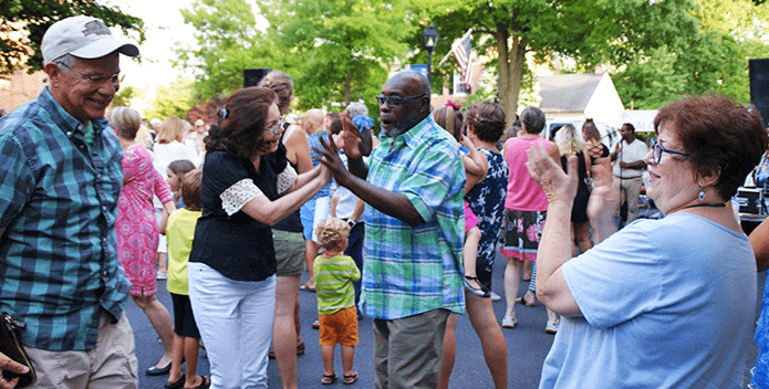 A couple dances in the street while others clap and gather infront of a music stage.