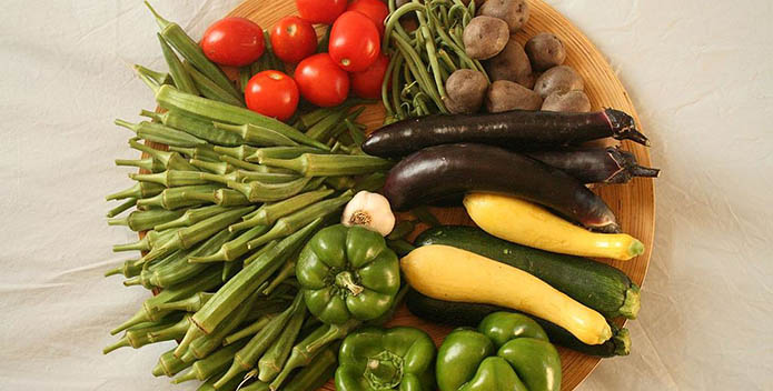 Photo of garlic, eggplant, yellow squash, zucchini, tomatoes, string beans, potatoes, green peppers, and okra arranged on a bamboo plate.