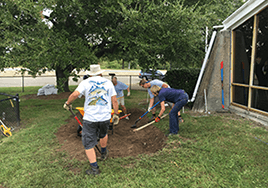 Four people work on digging the ground near a building to prepare for planting.