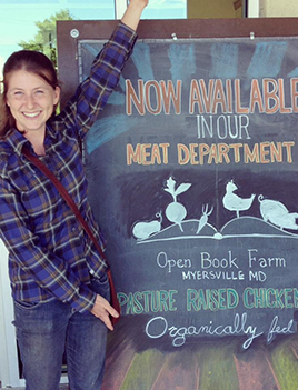 Proprietor of Open Book Farm, MK Barnet, stands in front of a sign adverting her products.