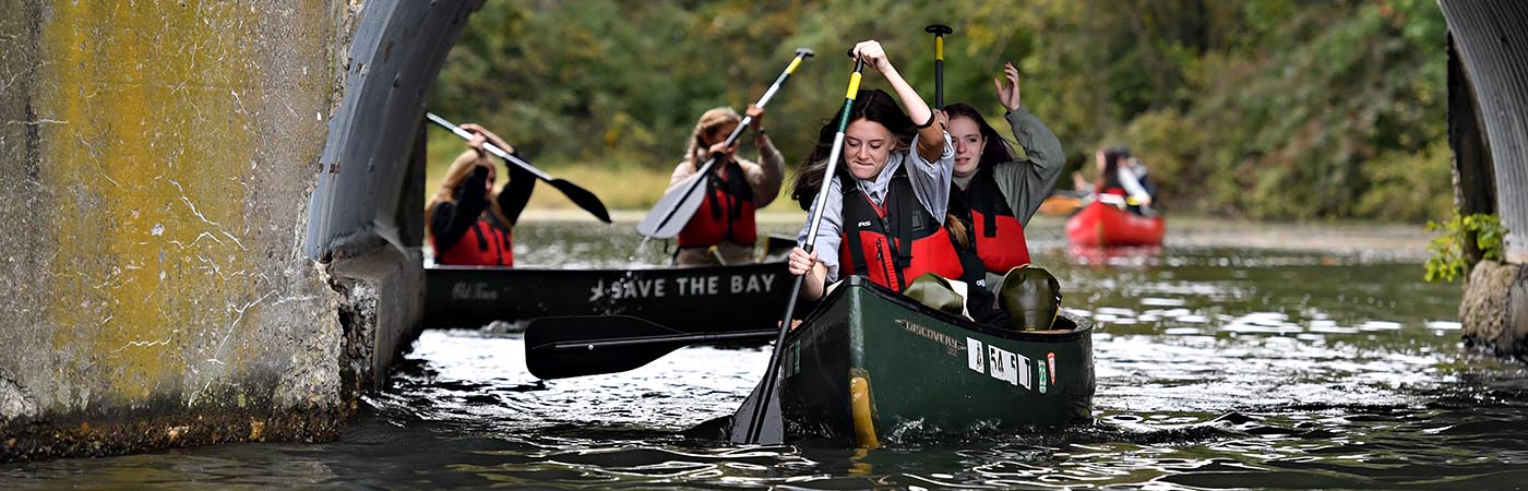 Students in red life jackets and green canoes paddle under a bridge.