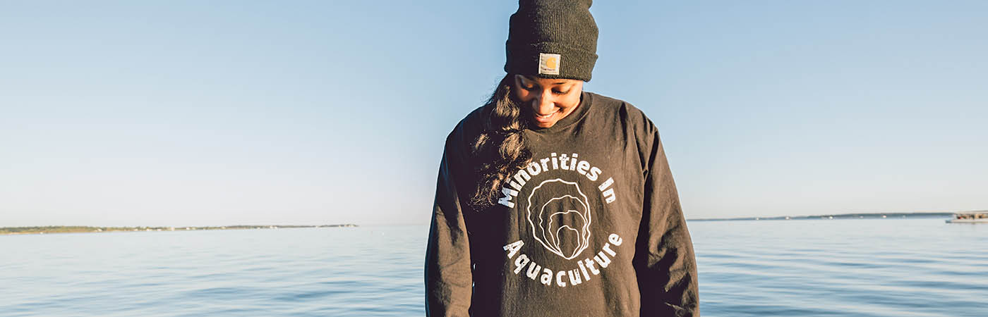 A young black woman stands in the Bay, wearing a sweatshirt that reads Minorities in Aquaculture.