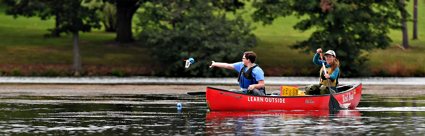 A student and teacher paddle in a red canoe, as the student tosses a blue floater in the water to collect samples.