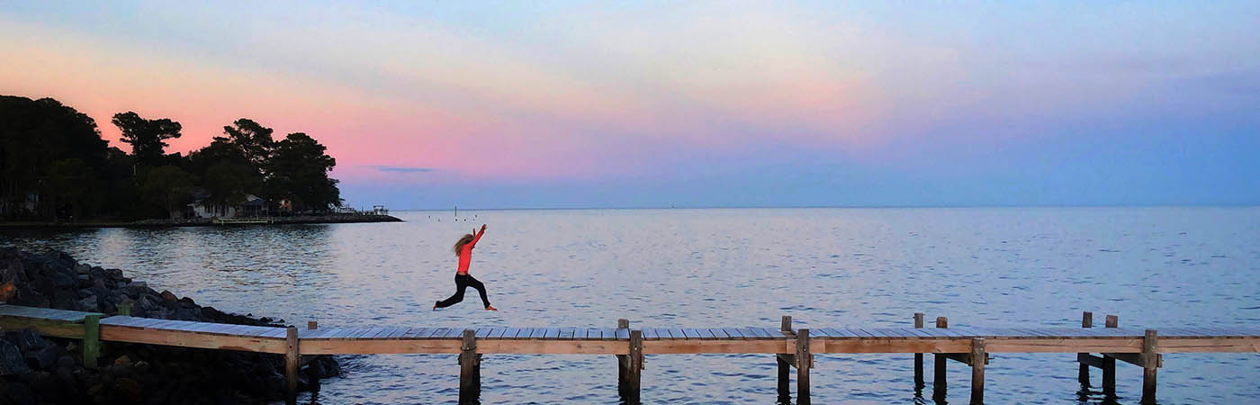 Woman in a red shirt jumping along a dock.
