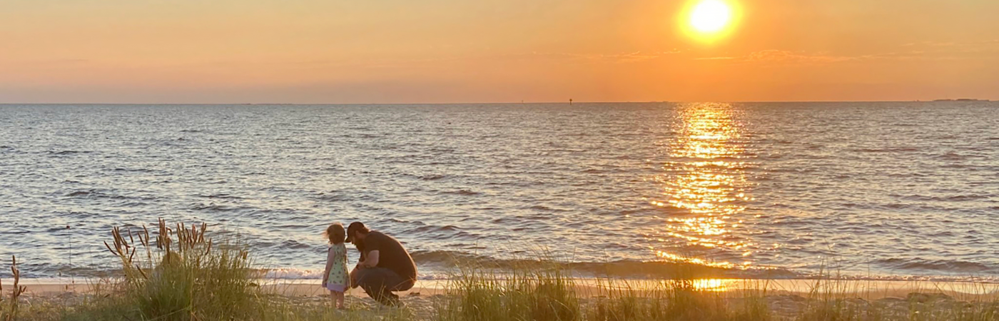 A father crouches down to talk to his young daughter on a beach in the early morning.