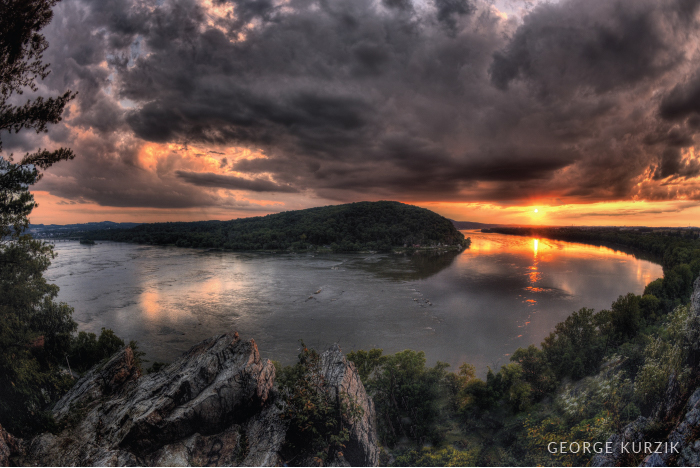An orange glow below dark clouds illuminates a towering rocky outcrop surrounded by water.