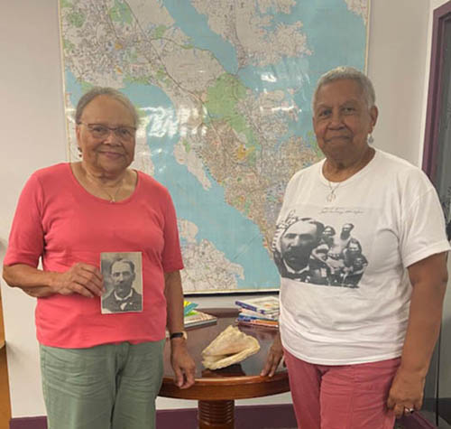 Two women standing by a table holding a shell with a wall map in the background.