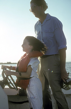 Young child, wearing a life vest steering a boat with an adult supervising.