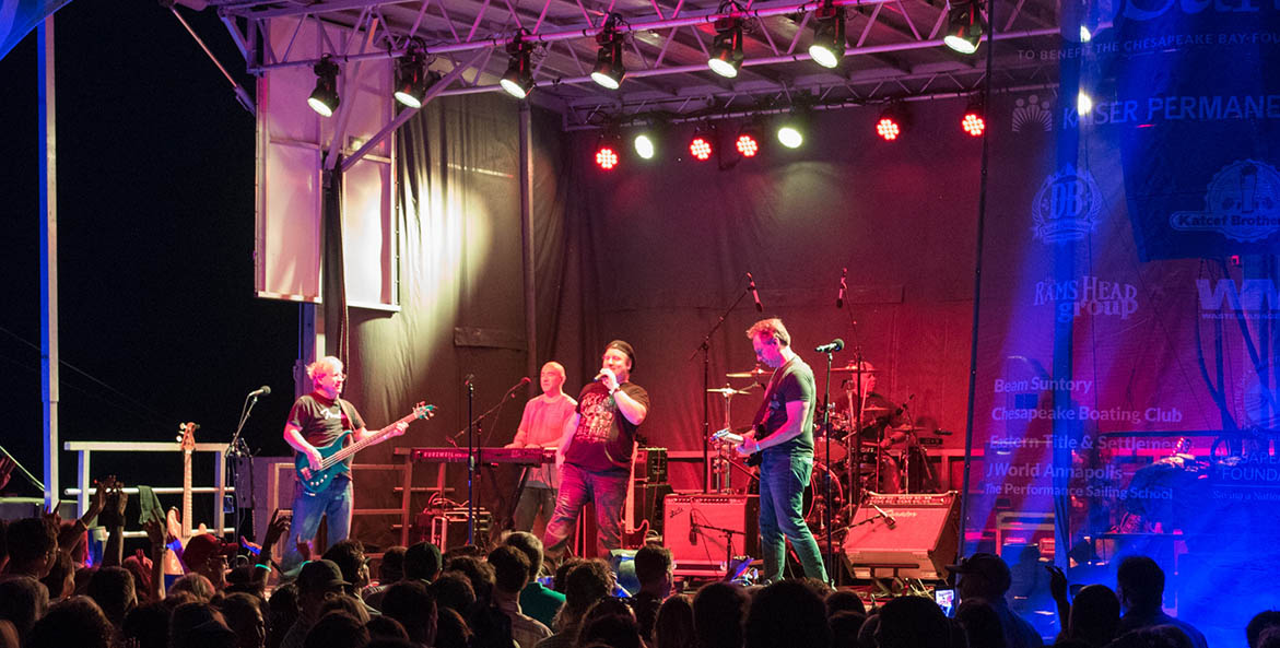 A band plays on an outdoor stage at night, the stage lights showing red and purple, as the audience stands in the foreground.