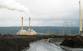 Steam rises from the stack of a power plant. A river flows alongside.