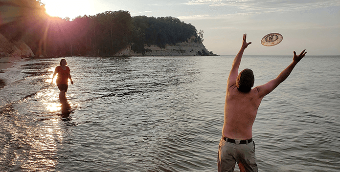 A man with his back to the camera reached for a frisbee while a woman looks on. Both are standing in the water as the sun sets in the background.