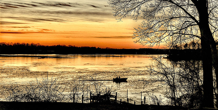 A shilouette of a boat with two figures rides along the shore and past a tree at sunrise.