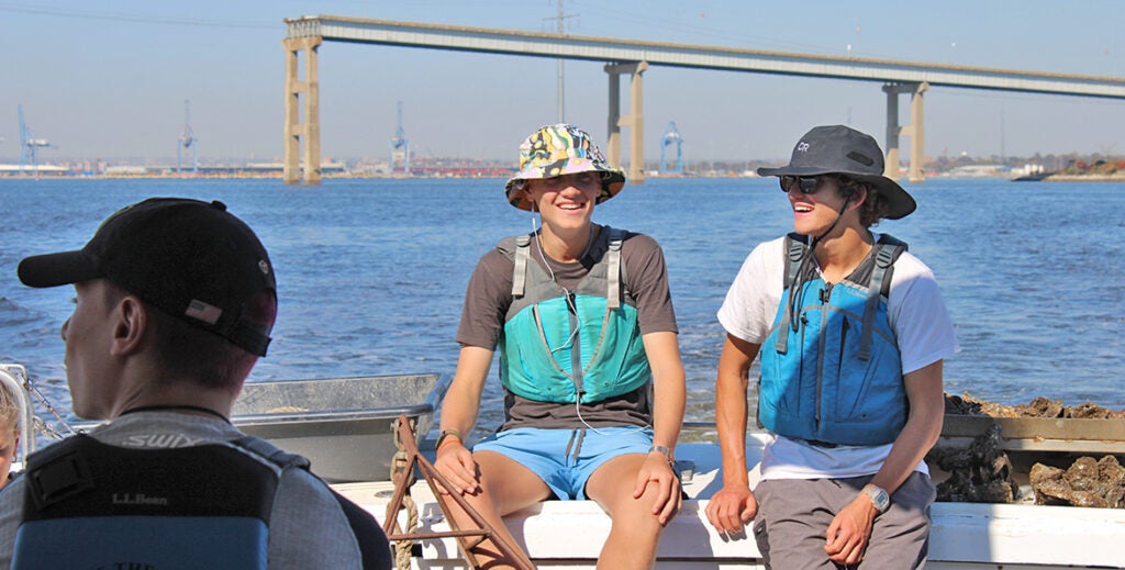 Two students sit at the stern of a boat with the remains of they Key Bridge in the background.