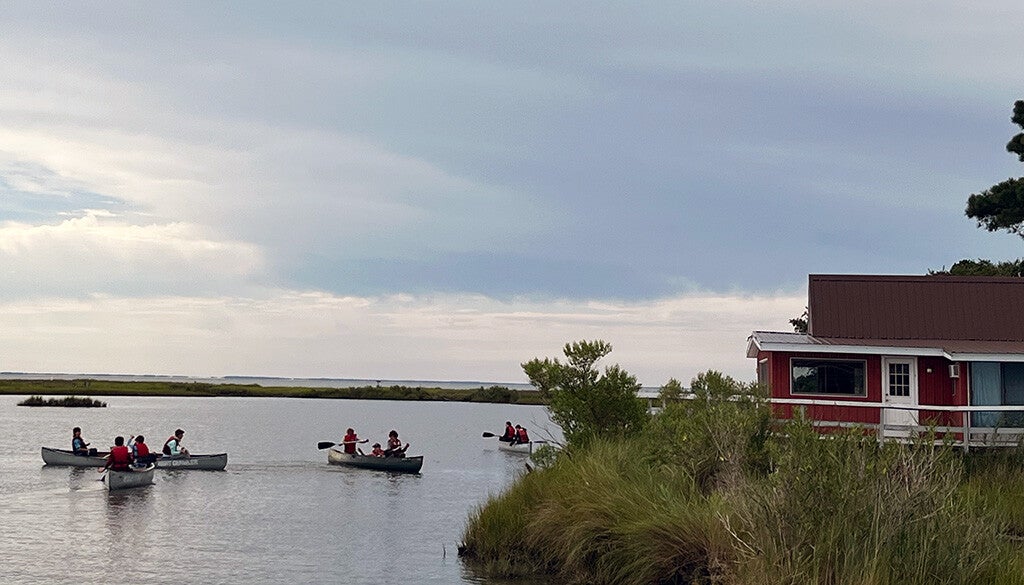 A group of canoeists paddles across a calm marsh under a broad, cloud-filled sky beside a red lakeside building.