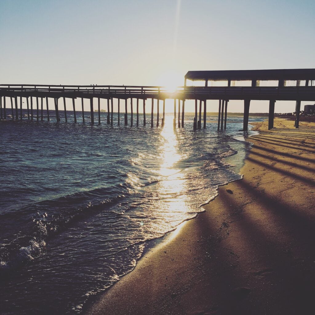 Sunset over a sandy beach with gentle waves and a long wooden pier extending into the water, casting long shadows across the shoreline as the sun reflects on the surface.