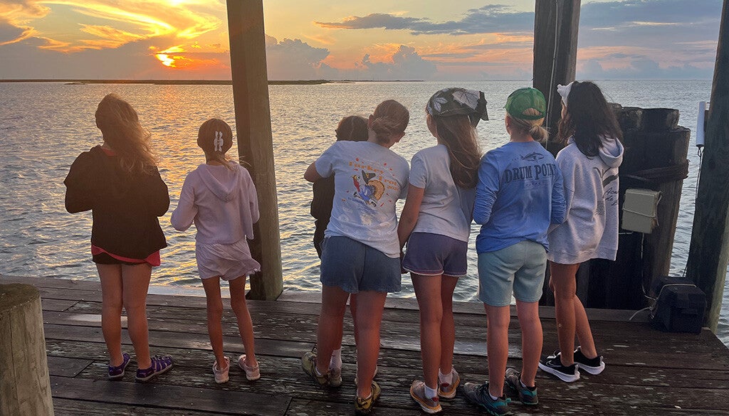 A group of young students watches the sun melt into the horizon from the dock, soaking in the golden hush of a summer evening.