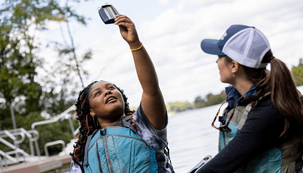 Two people wearing life vests are standing near a dock by the water. One person is holding a small device high in the air. Trees and a cloudy sky are visible in the background.