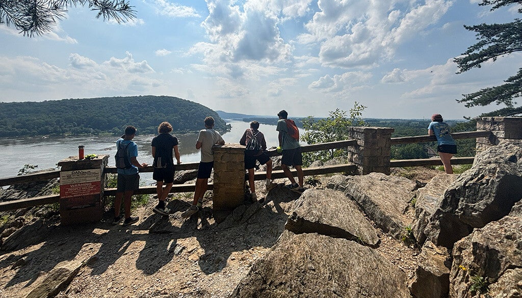 A group of hikers pauses at a rocky overlook to take in the river valley and dramatic cloud-streaked sky.