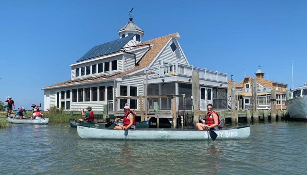 Two people in red life jackets paddle a canoe labeled “SALTY BIRD” past sunlit waterfront houses under a clear blue sky.