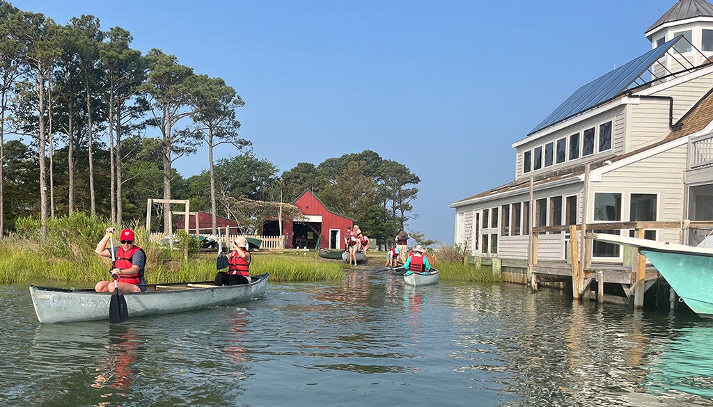 A group of paddlers in red life jackets glide past a waterside boathouse under a clear blue sky.