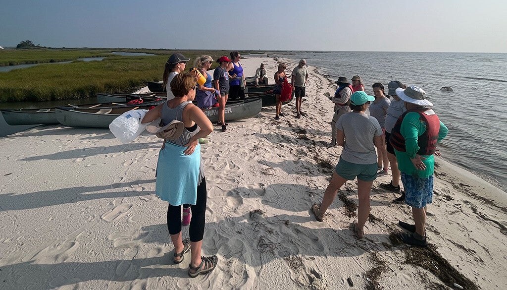 A group of paddlers stands on a narrow sandy spit next to several canoes, preparing to head out into the calm coastal waters under a clear blue sky.