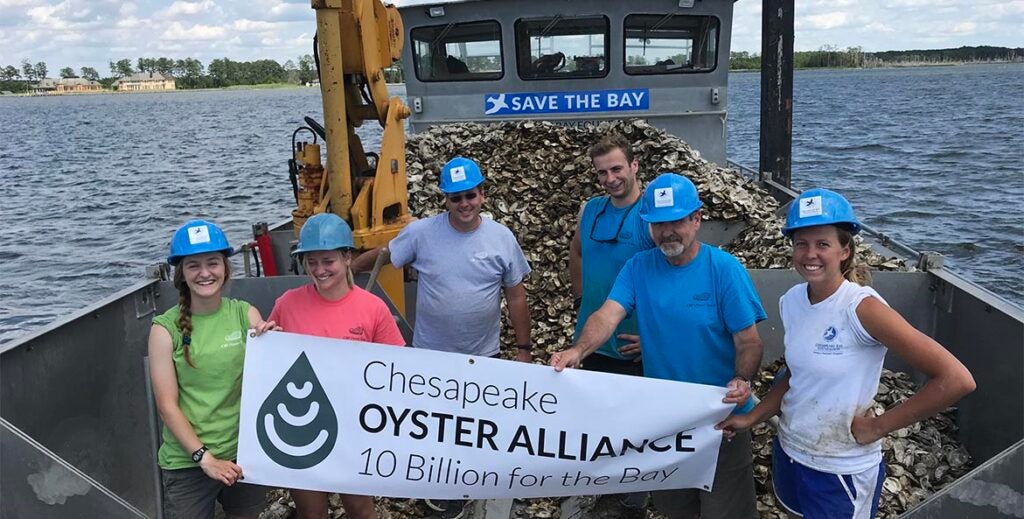 Five volunteers wearing blue hard hats hold a Chesapeake Oyster Alliance banner on a barge loaded with oyster shells