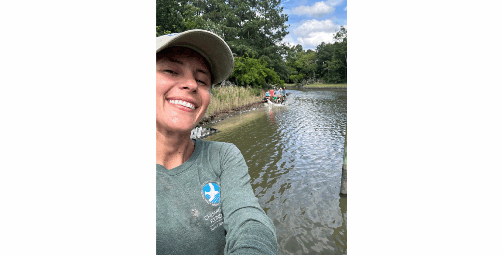 A smiling woman in a cap and Chesapeake Bay Foundation shirt takes a selfie beside water with a group of people visible along the shore in the background.