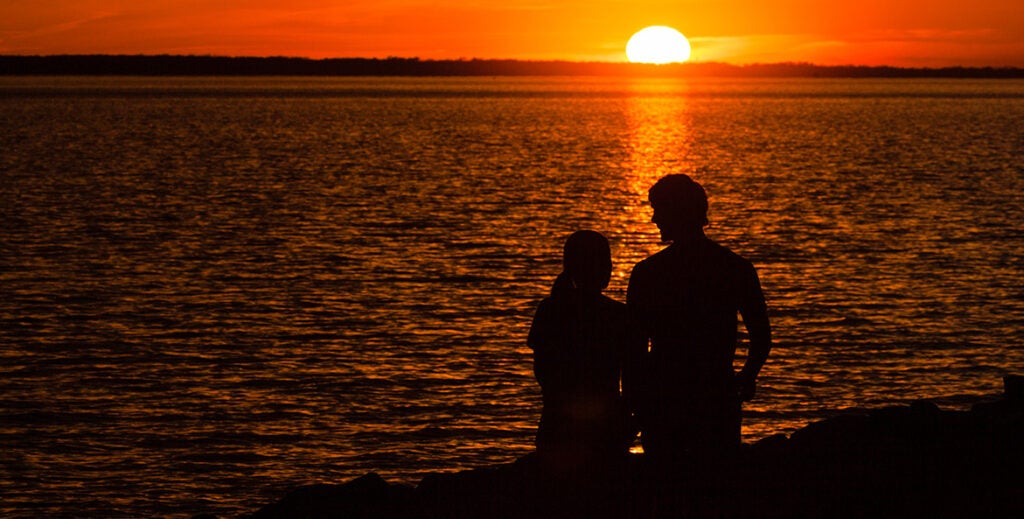 Two people watch the sun set over a river.