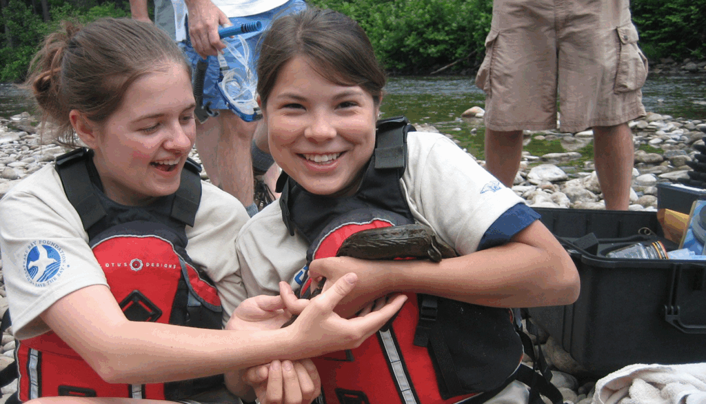 Two people wearing life jackets sit on a rocky riverbank, gently holding an Eastern hellbender. Other individuals stand nearby, and equipment cases are visible in the background near the water.