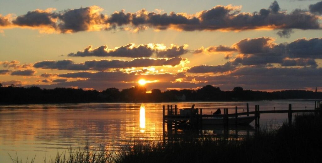 Sunrise over the Chesapeake Bay with dramatic clouds, golden light reflecting on calm water, and a silhouetted dock in the foreground.