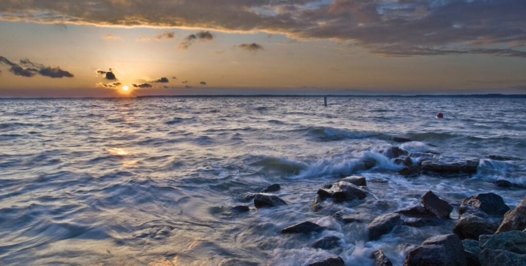 Sunset over a body of water with waves breaking over rocks and golden light streaming through clouds on the horizon.