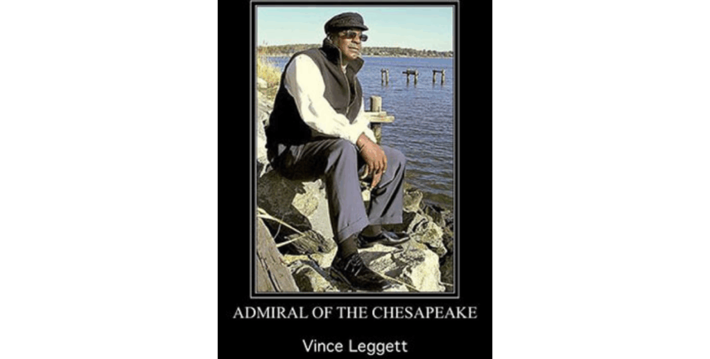 ince Leggett, "Admiral of the Chesapeake," sits on rocks beside the bay wearing sunglasses and a vest.