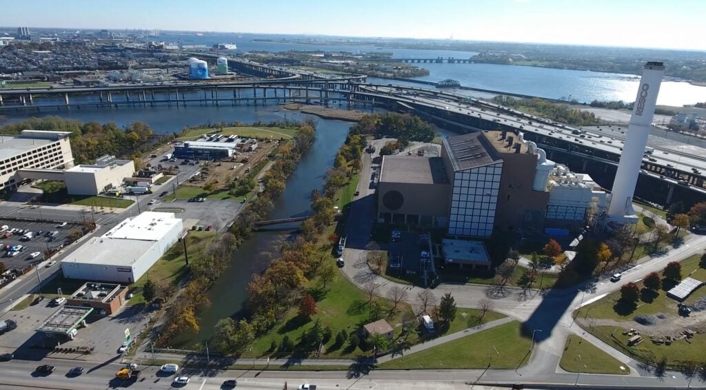 Aerial view of an industrial facility near a waterway, with surrounding roads, bridges, and buildings; a large smokestack and green spaces are visible, and a wide river extends toward the horizon under clear skies.