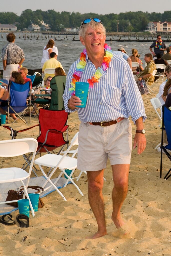 A gray-haired man in a blue and white striped shirt and khaki shorts and flowered lei holds a cup while standing on the beach, surrounded by people relaxing in chairs.