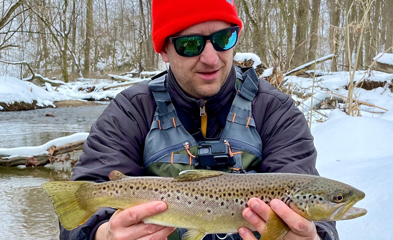 A man dressed for cold weather is kneeling next to a shallow, partially frozen stream in a snowy woodland holding a brown trout.