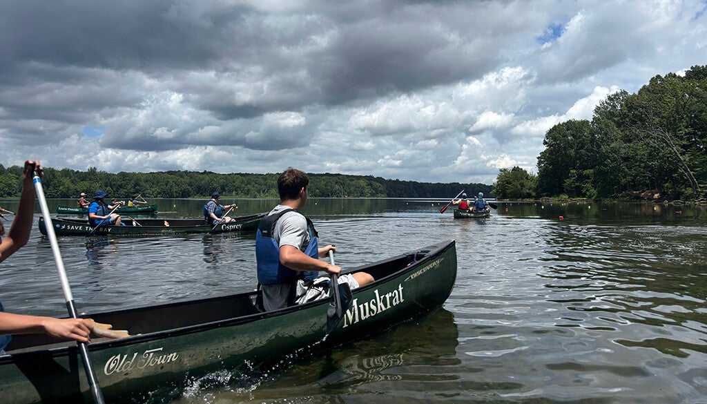 A group of canoeists paddle across a calm lake beneath a dramatic, cloud-filled sky.
