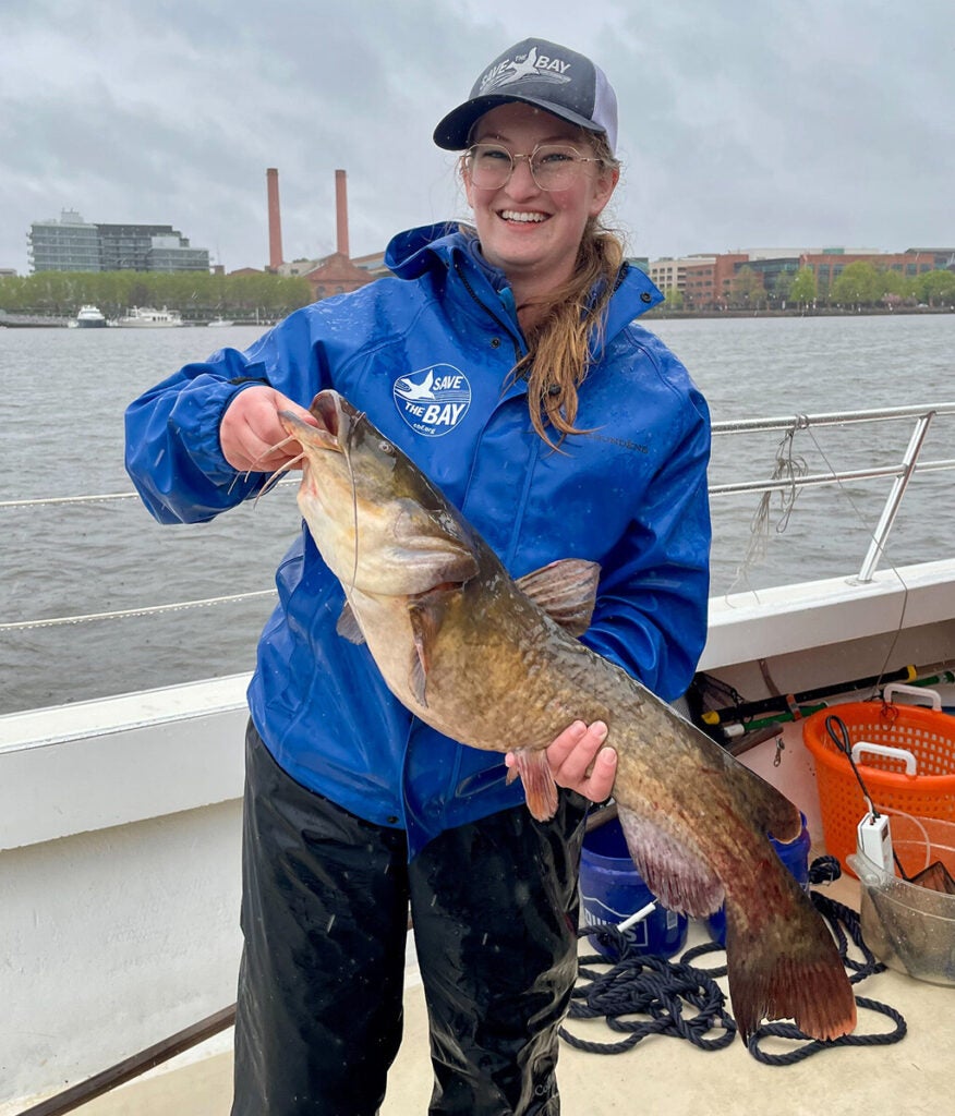 A smiling young woman stands on the deck of a boat holding a large catfish.