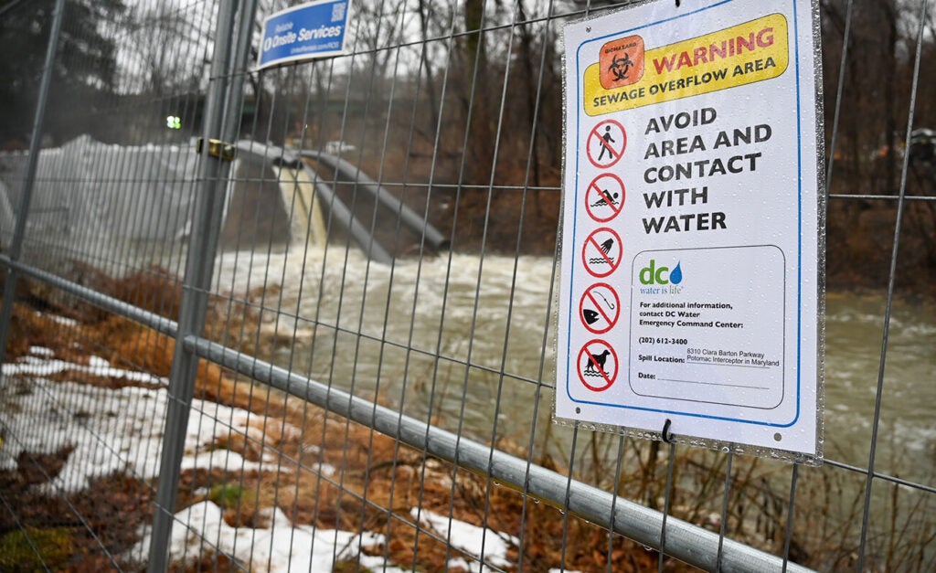 Sewage filled water spills from pipes into a river behind a fence bearing a warning sign.