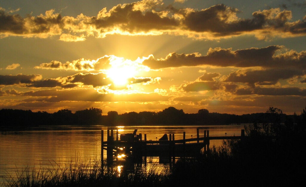 Silhouetted dock on the Chesapeake Bay at sunrise with golden light breaking through dramatic clouds and reflecting on calm water