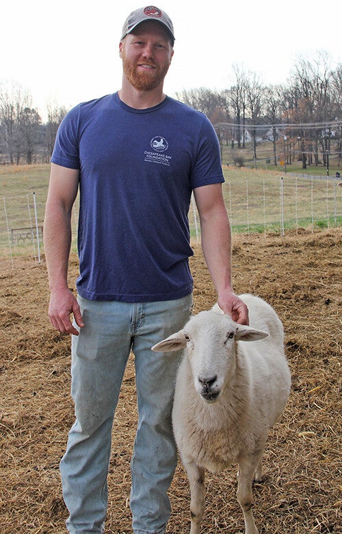 Alex Outten, in a gray ball cap and blue CBF t-shirt, stands in a field with a sheep.