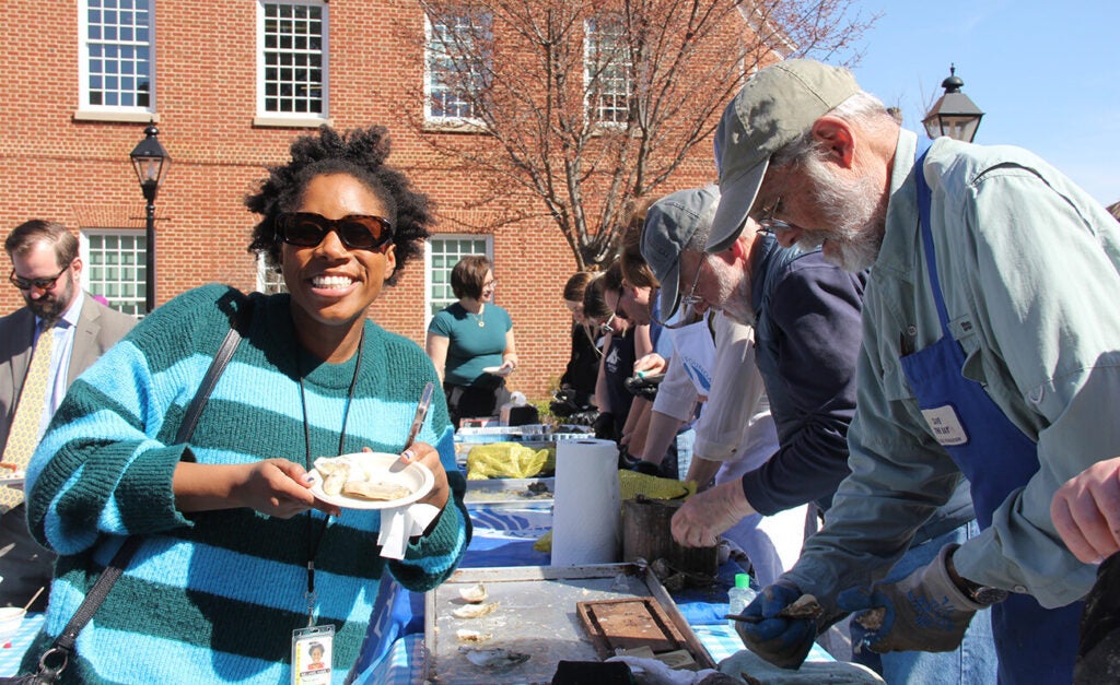 A woman holds a plat with oysters while standing in front of a table of people shucking oysters.