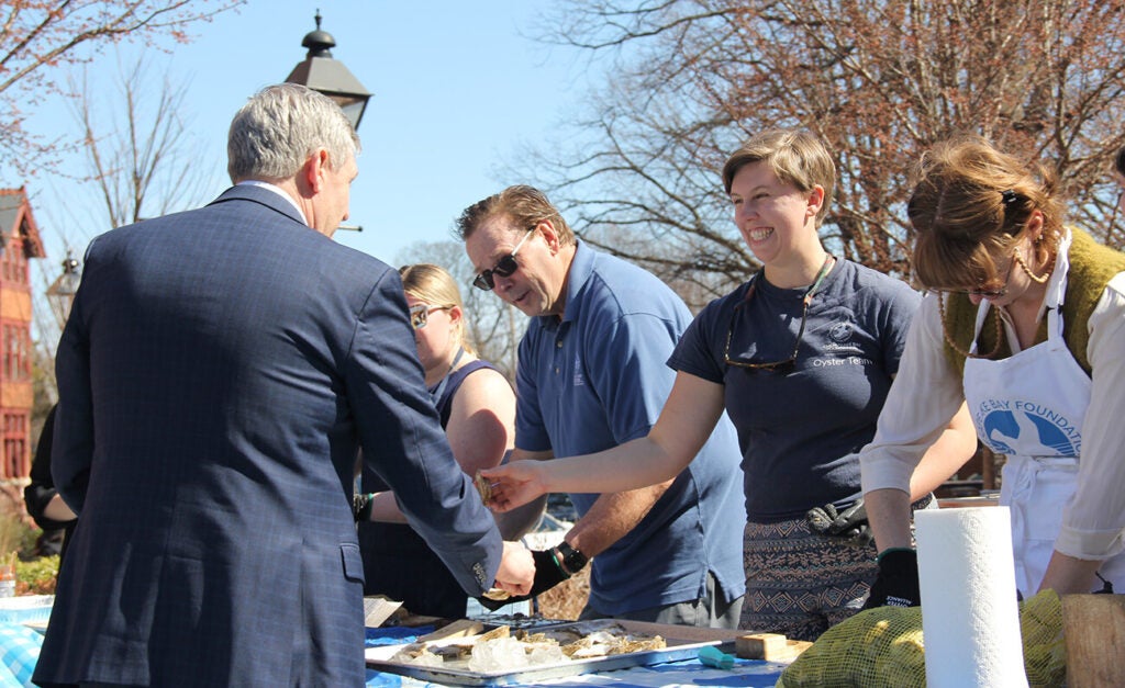 A smiling woman hands a man a newly shucked oyster while others stand next to her at a table shucking more oysters.