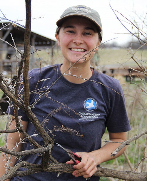 Kayla Brennan, in a gray Rocky Mountain cap and blue CBF t-shirt stands behind a bush with a pair of pruning shears in her hand.