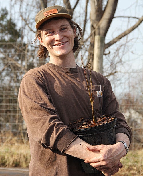 Will Grinnell, in a brown cap and brown shirt, holds a tree sapling.