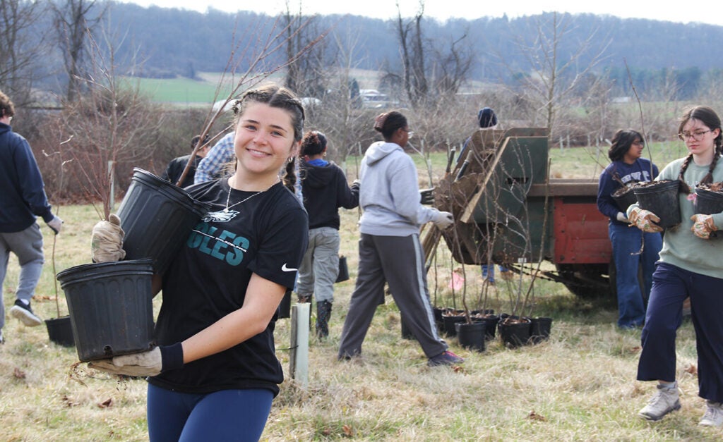 A group of young people plant saplings in a field while a smiling woman carries two potted young trees.