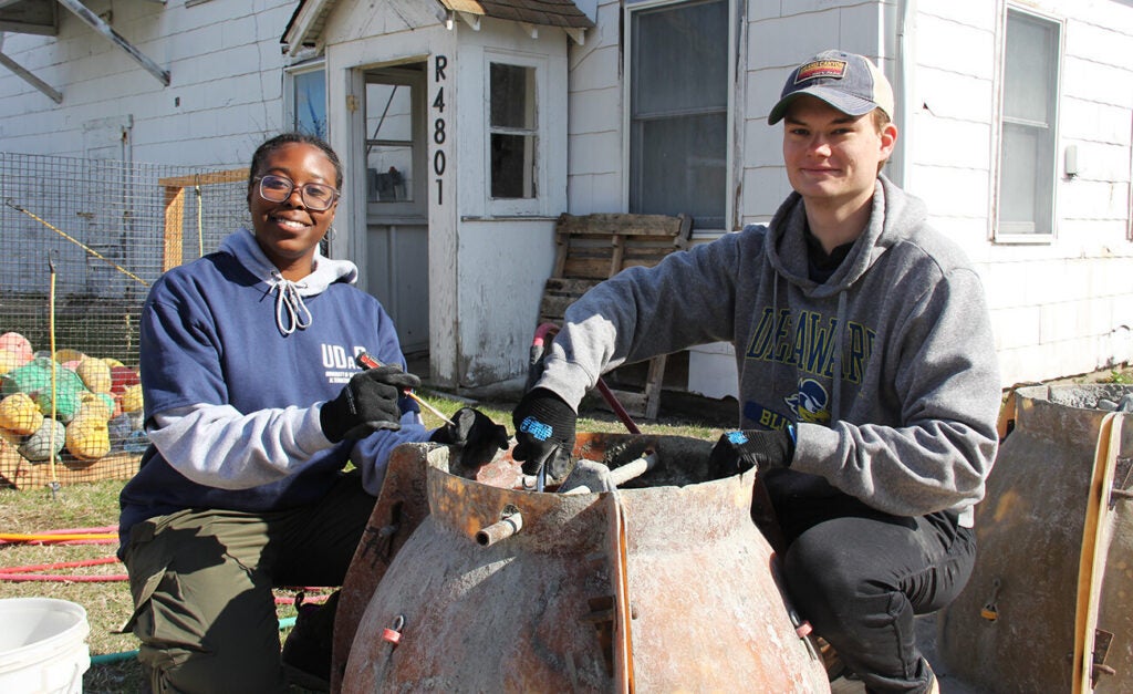 A young woman and young man kneel and smile while working on a concrete form in front of a white house.